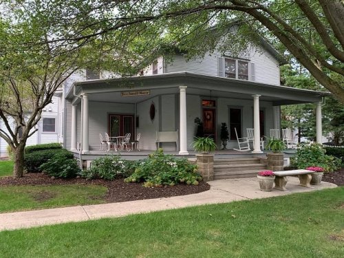 A charming two-story house with a porch, surrounded by greenery and flower beds, featuring rocking chairs and a stone bench.