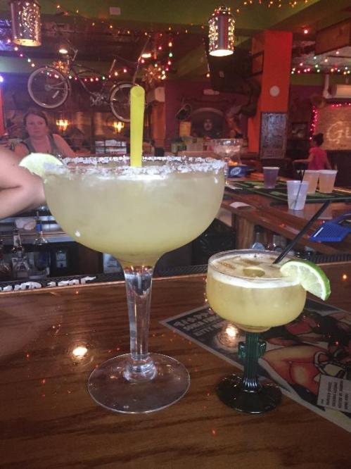 A large margarita in a tall glass with a lime wedge and a smaller margarita in a short glass on a bar counter.