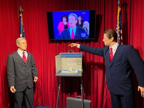 Two lifelike wax figures of politicians stand in front of a TV screen, with a voting box and flags in the background.