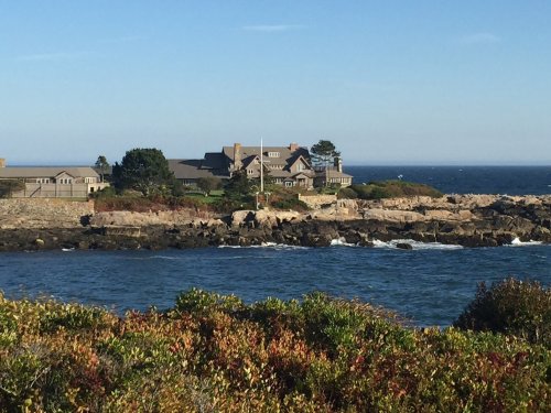 A coastal mansion overlooks the ocean, surrounded by rocky shores and greenery under a clear blue sky.
