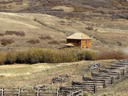 A rustic wooden house sits on a hillside, surrounded by sparse vegetation and a wooden fence in a dry landscape.
