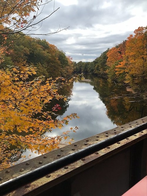 A serene river reflects colorful autumn foliage under a cloudy sky, viewed from a bridge.