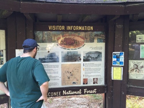 A person stands in front of a visitor information sign at the Conoree National Forest, reading about local history.