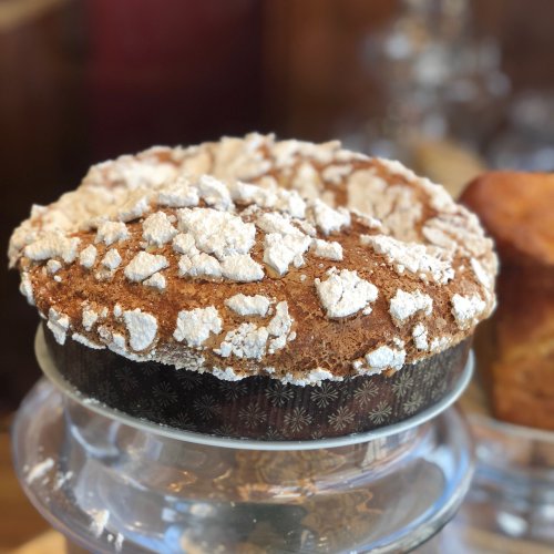 A freshly baked cake with a textured, powdered sugar topping, displayed on a glass cake stand.