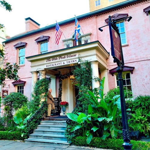 A historic pink building with flags, featuring a welcoming entrance surrounded by greenery and a sign for a restaurant.