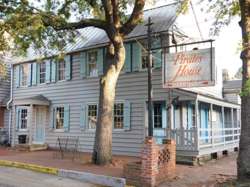 Historic building with blue shutters and a sign reading "Pirates House," surrounded by trees and brick pavement.