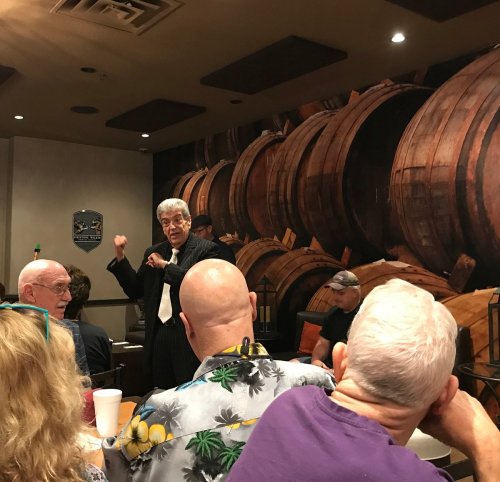 A speaker gestures while addressing an audience in a room decorated with large wooden barrels.