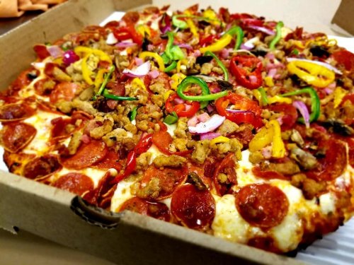 A close-up of a pizza topped with pepperoni, sausage, colorful peppers, and onions in a takeout box.