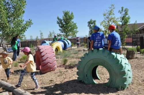 Children play on colorful tire structures in a sunny outdoor setting, surrounded by trees and grass.