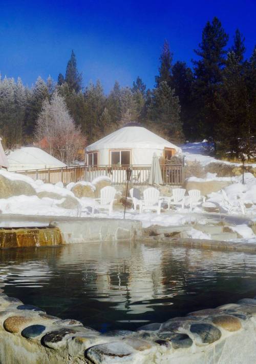 A serene winter scene featuring a hot spring pool, white chairs, and a yurt surrounded by snow-covered trees.