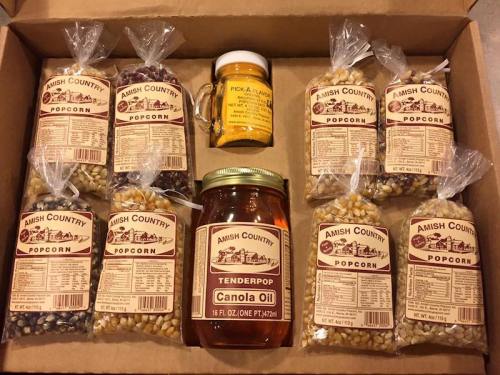 A box containing various popcorn bags, a jar of canola oil, and a small jar of seasoning, all labeled Amish Country.