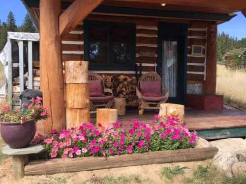 A cozy log cabin porch with two chairs, stacked firewood, and vibrant pink flowers in a pot.