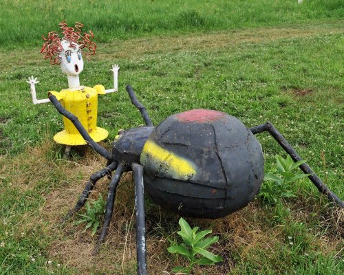 A whimsical metal sculpture of a girl with curly hair stands beside a large, painted spider in a grassy field.