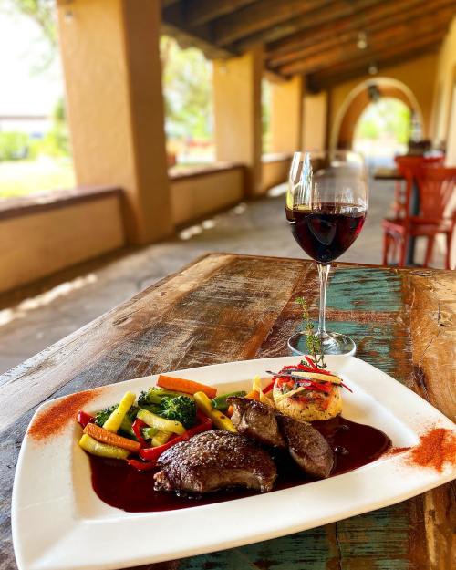 A plate of grilled meat with vegetables and a glass of red wine on a rustic wooden table in a warm, inviting setting.
