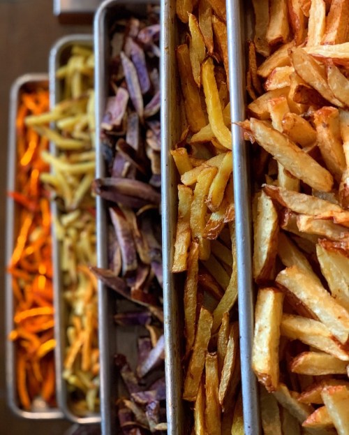 A variety of freshly cooked fries in metal trays, including golden, purple, and sweet potato fries.
