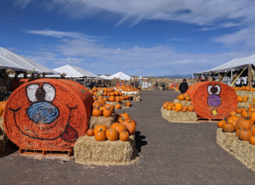 Two large hay bales with cartoon faces, surrounded by pumpkins, under a blue sky with white clouds.