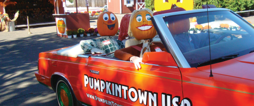 Two pumpkin-headed figures sit in a convertible car, promoting Pumpkintown USA at a festive outdoor location.