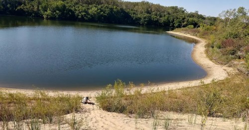 A serene lake surrounded by greenery and a sandy shore, with a person sitting near the water's edge.