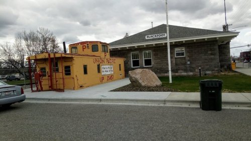 A yellow train caboose next to a stone train station, with a large rock and trash can in the foreground.