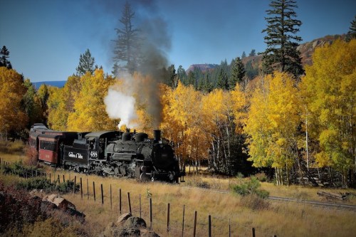 A vintage steam train chugs through a vibrant autumn landscape with golden trees and a clear blue sky.