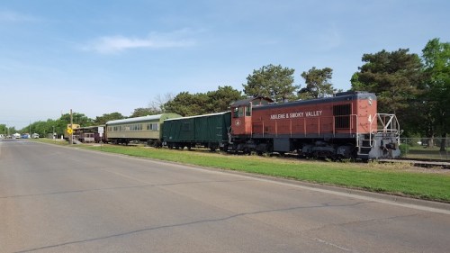 A red train engine and several cars parked beside a road, surrounded by green trees and clear blue skies.
