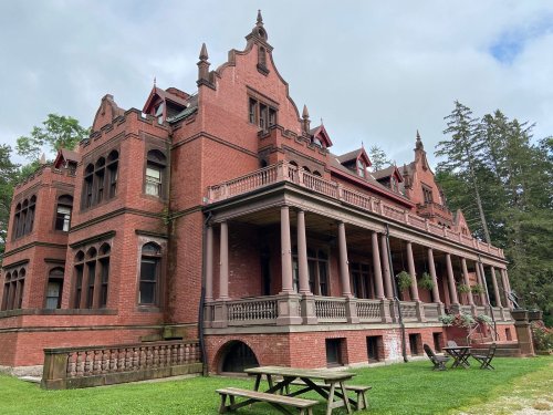 A large, red brick mansion with ornate architecture, featuring a wraparound porch and surrounded by greenery.