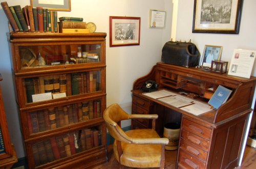 A vintage wooden desk with a chair, bookshelves filled with books, and framed pictures on the wall in a cozy room.
