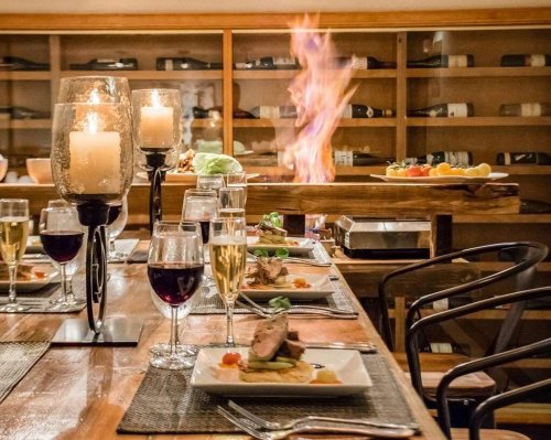 A beautifully set dining table with candles, wine glasses, and a flaming dish in the background, creating an elegant atmosphere.