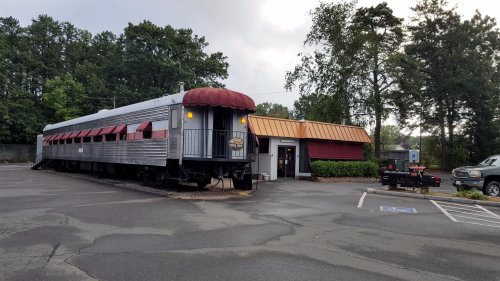 A vintage train car converted into a restaurant, parked beside a building in a tree-lined area.