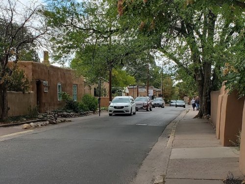 A quiet street lined with adobe buildings, trees, and parked cars, with pedestrians walking in the distance.
