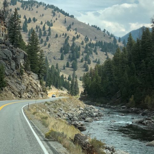 A winding road alongside a river, surrounded by mountains and dense trees under a cloudy sky.