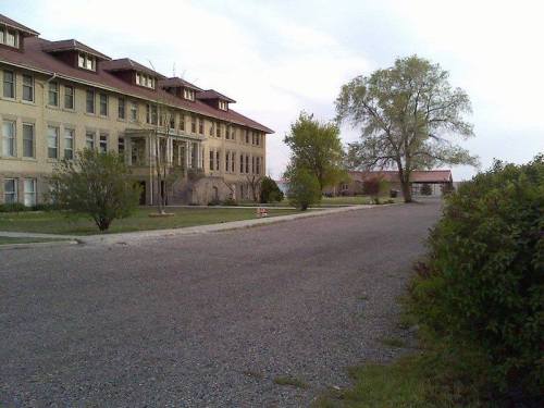 Abandoned building with a large porch, surrounded by trees and a gravel road, under a cloudy sky.