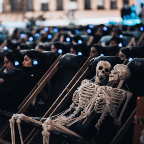A group of people in headphones relaxes in lounge chairs, with a skeleton sitting among them.