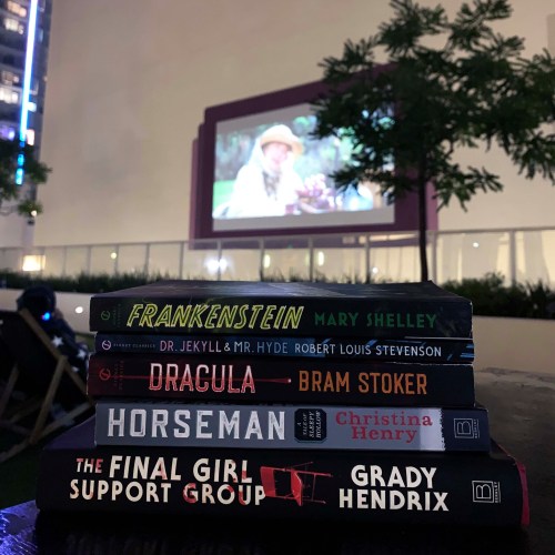 A stack of books including "Frankenstein," "Dracula," and "The Final Girl Support Group," with an outdoor movie screen in the background.
