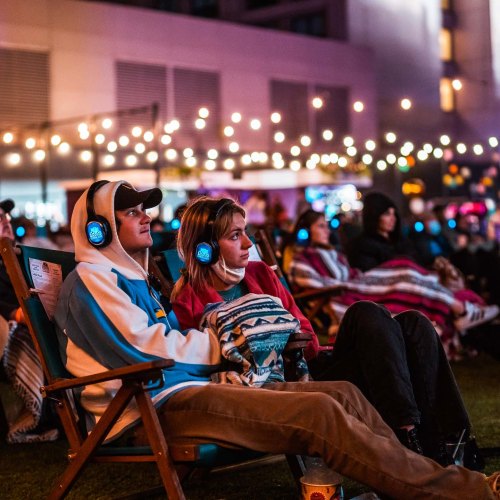 Two people in outdoor chairs wearing headphones, wrapped in blankets, enjoying a nighttime event with string lights in the background.