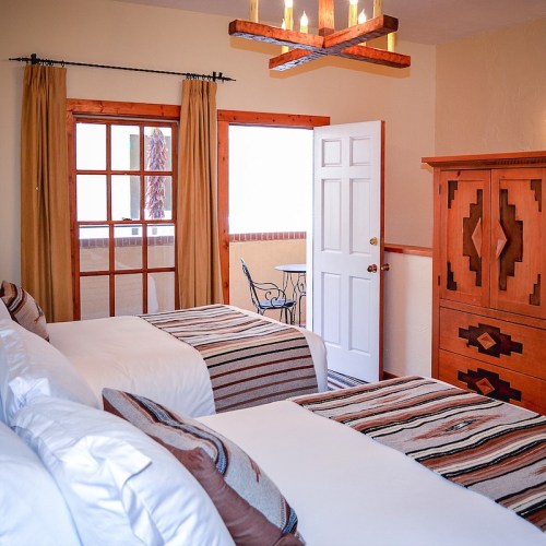 Cozy bedroom with two beds, wooden furniture, and warm lighting from a chandelier. Windows with curtains add natural light.