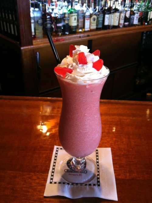 A tall glass of pink smoothie topped with whipped cream and strawberries, set on a bar counter with bottles in the background.