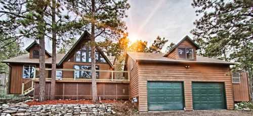 A wooden house with large windows, surrounded by trees, featuring a two-car garage and a deck. Sunlight peeks through the trees.