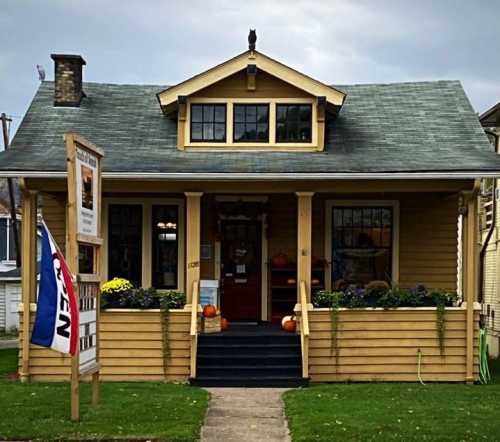 A charming yellow house with a porch, decorated with pumpkins and flowers, under a cloudy sky. A "Open" sign is visible.