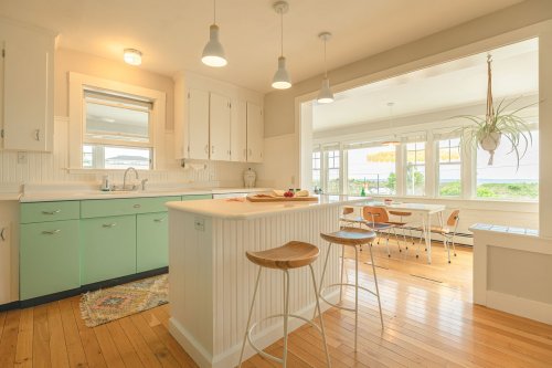 Bright kitchen with mint green cabinets, wooden floors, and a dining area featuring large windows and a view.