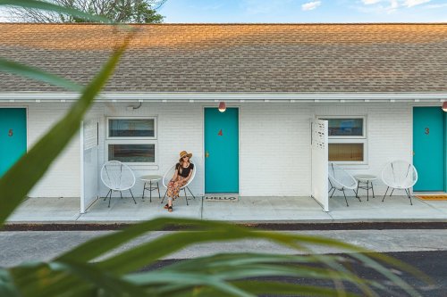 A woman sits outside a motel room with turquoise doors, surrounded by greenery and a sunny sky.