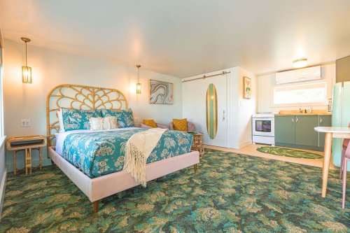 Cozy bedroom with a floral bedspread, green carpet, and modern kitchen area, featuring natural light and stylish decor.