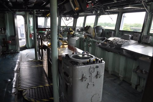 Interior of a ship's bridge, featuring control panels, steering wheel, and large windows with a view outside.