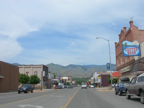 A small town street lined with shops, including a coffee shop, with mountains in the background under a cloudy sky.
