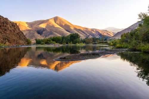 Serene river scene reflecting golden mountains at sunset, surrounded by lush greenery and calm waters.