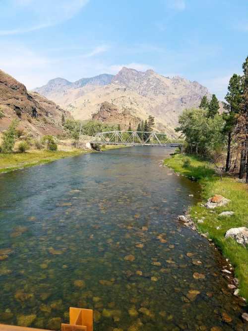 A serene river flows through a mountainous landscape, with green banks and a bridge in the distance under a clear blue sky.