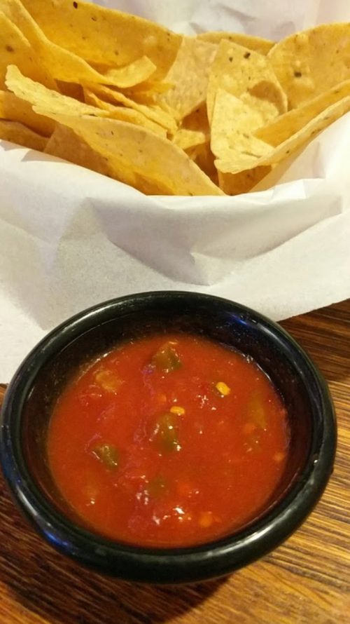 A bowl of red salsa sits in front of a basket of crispy tortilla chips.