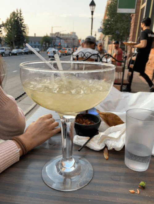 A large margarita glass filled with ice sits on a table, with people dining in the background on a sunny day.