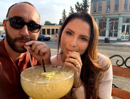 A couple enjoys a large cocktail outdoors, smiling and sipping through straws, with a sunny street scene in the background.