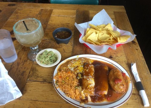 A plate of enchiladas with rice, a side of guacamole, tortilla chips, and a margarita on a wooden table.